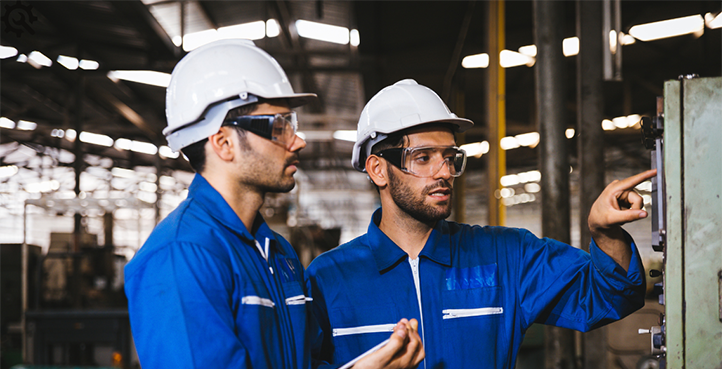 Zwei Arbeiter mit weißen Helmen und blauen Overalls bei einer Sicherheitsprüfung an einer Maschine in einer Industriehalle